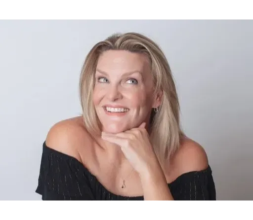 Fiona O’brien The image is a professional portrait of a woman named Fiona O'Brien. She has blonde hair, parted slightly off-center, and a friendly, engaging smile. Fiona's pose is relaxed and confident, with her chin resting on her right hand and her elbow on the table or surface. She is wearing an off-the-shoulder black top, revealing her shoulders and collarbones. The background is a plain, light color, which keeps the focus on her face and expression. Fiona's makeup is subtle and enhances her natural features, adding to the polished look of the portrait.