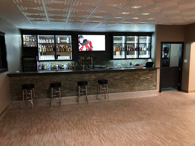 This image features a modern bar setup with a compact yet stylish design. The stone-faced bar counter is paired with sleek metal stools, creating a clean and minimalist aesthetic. Behind the bar, illuminated shelves display a variety of beverages, with a central TV screen adding entertainment value. The neutral-toned flooring and ceiling provide a balanced and welcoming ambiance, ideal for casual events or social gatherings.